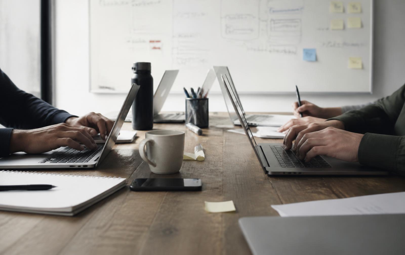 Two people working side by side on laptops at a wooden table in front of a whiteboard covered in diagrams